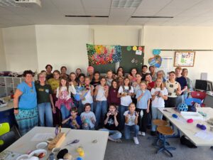 Eine Gruppe von Schülern und Lehrern posiert lächelnd in einem hellen Klassenzimmer in Bretten, das mit bunten Bastelarbeiten geschmückt ist. Auf den Tischen liegen Malutensilien, Garn und Bastelmaterial. Einige Schüler halten selbstgemachte Bastelarbeiten in den Händen, und alle wirken glücklich.