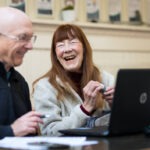 Ein älterer Mann und eine Frau sitzen zusammen an einem Tisch, lächeln und lachen, während sie einen Laptop benutzen. Die Frau hält eine Computermaus, und beide scheinen einen freundlichen, entspannten Moment in einem hellen Innenraum zu genießen.