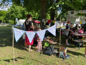 Bei einer Gemeinschaftsveranstaltung in Augsburg versammeln sich die Menschen im Freien unter Bäumen. Auf den Wimpeln steht „Omas for Future“. An den Tischen werden Bastelaktivitäten angeboten, und im Hintergrund hängen bunte Banner, während Kinder und Erwachsene im sonnigen Park miteinander spielen.