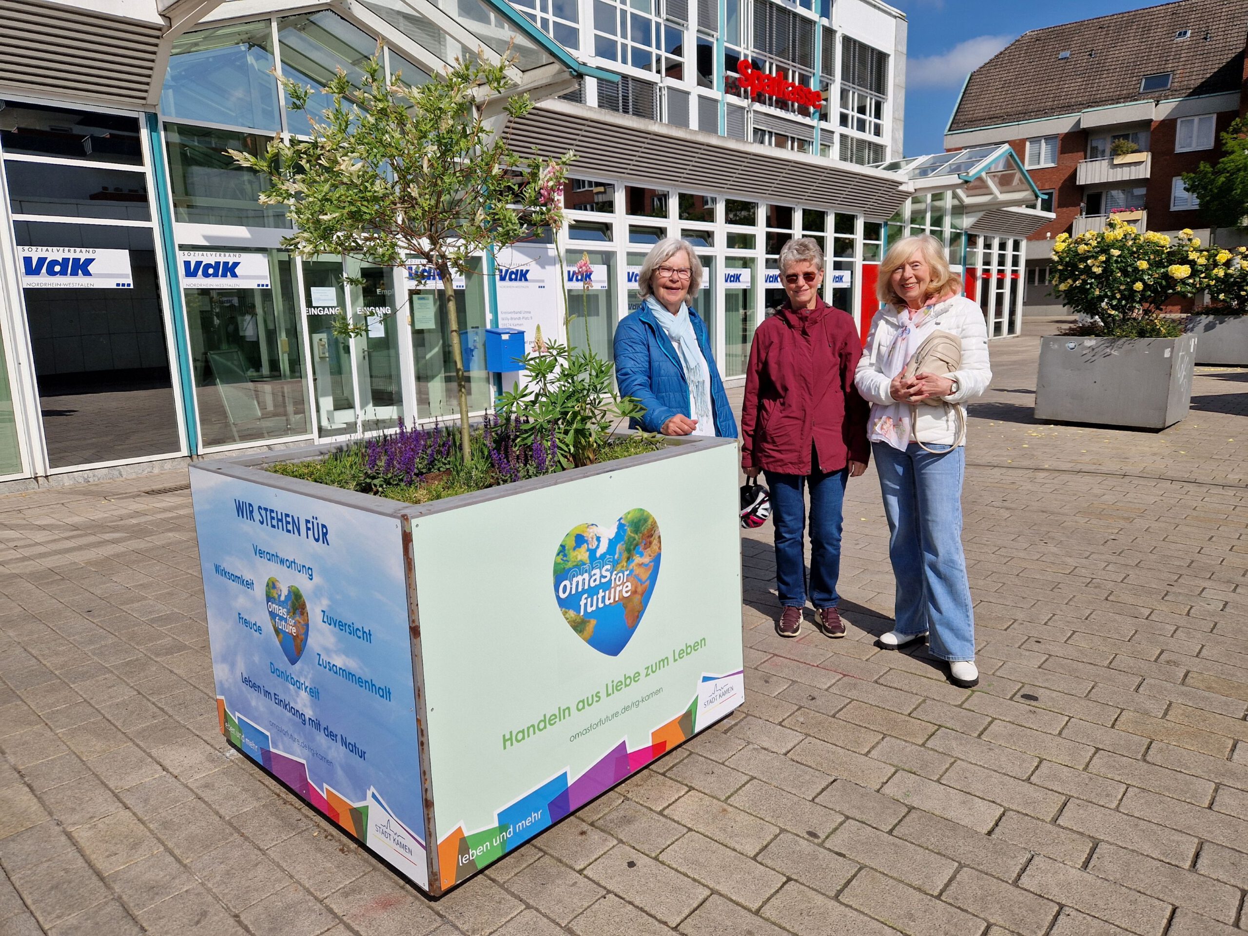 Drei ältere Frauen stehen neben einem großen Pflanzkübel mit Grünzeug auf einem gepflasterten Platz vor einem Gebäude. Auf dem Pflanzkübel ist ein buntes Schild mit dem Hinweis auf Umweltschutzmaßnahmen angebracht.
