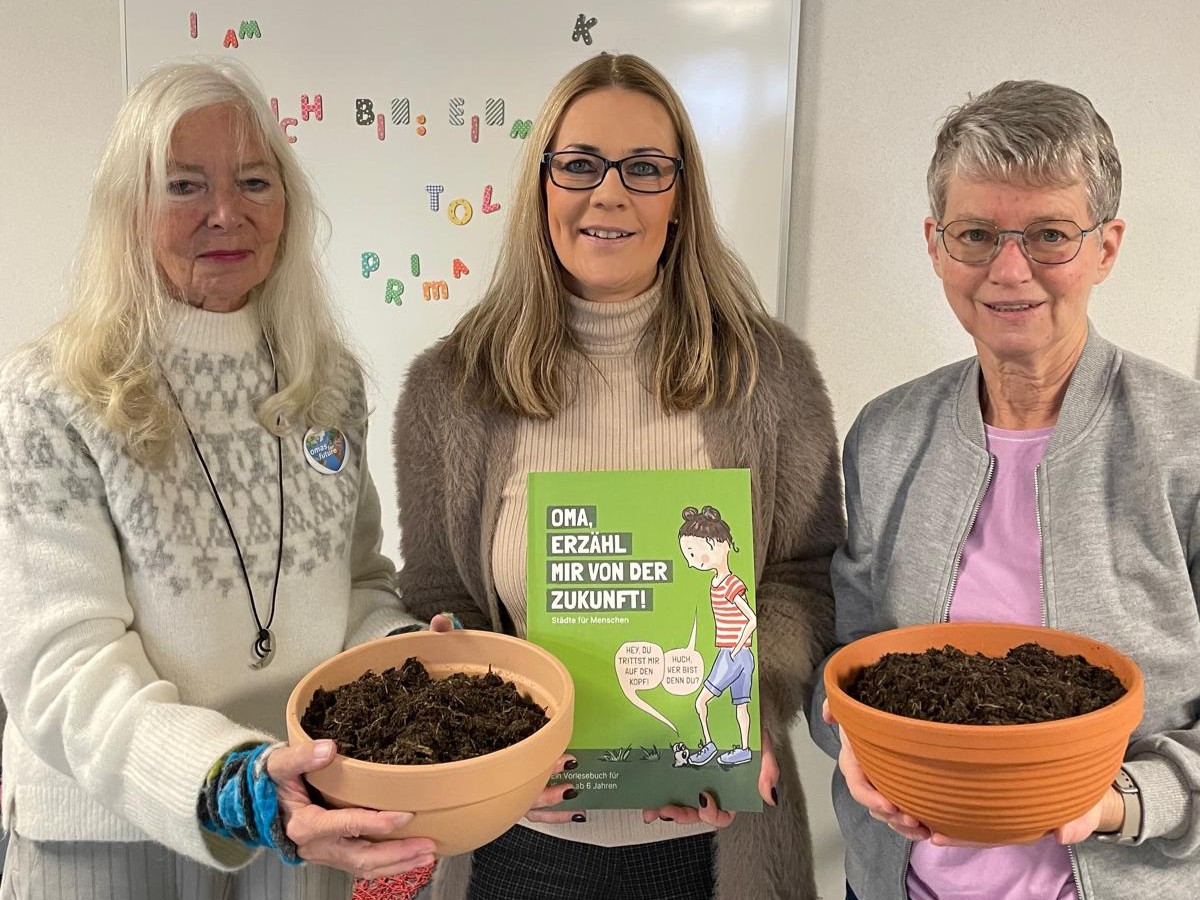 Drei Frauen stehen in einem Haus, zwei halten Schalen mit Erde. Die Frau in der Mitte hält ein deutschsprachiges Buch mit dem Titel Oma, erzähl mir von der Zukunft! Sie lächeln, und an der Wand hinter ihnen ist ein buntes Schild angebracht.