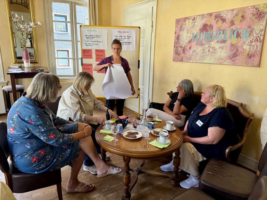 Fünf Frauen sitzen um einen runden Holztisch mit Kaffeetassen, Gebäck und Papieren. Eine Frau steht an einer Tafel mit Notizen, hält ein großes leeres Blatt in der Hand und leitet eine Diskussion in einem gemütlichen, gut beleuchteten Raum.
