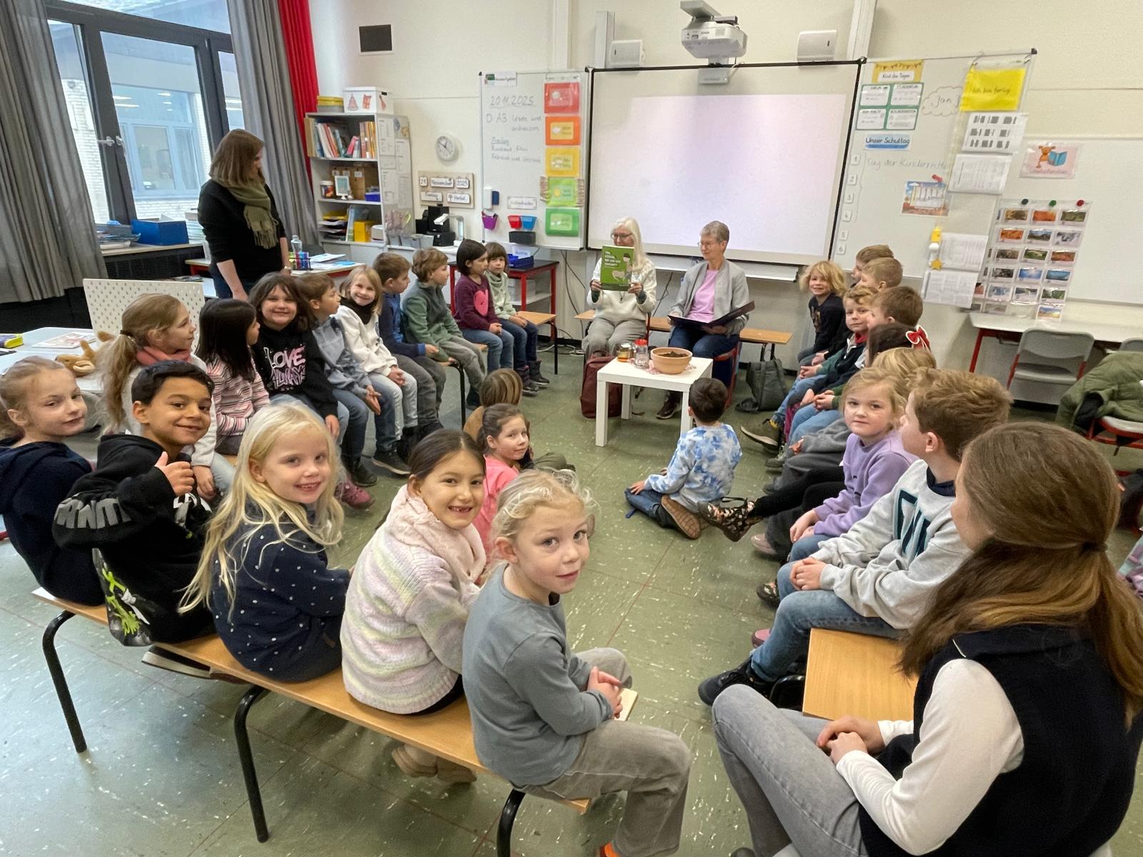 Ein Klassenzimmer mit kleinen Kindern, die in einem Kreis auf Bänken und Stühlen sitzen, lächeln und in die Kamera schauen. Vorne sitzen zwei Erwachsene, und im Hintergrund sind Unterrichtsmaterialien und ein Projektor zu sehen.