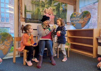 Eine ältere Frau sitzt auf einem Stuhl und liest zwei kleinen Mädchen in einem gemütlichen Raum vor, der mit herzförmigen Omas for Future-Schildern und einem großen Poster dekoriert ist. Die Kinder lächeln und sind beschäftigt, sie halten bunte Spielsachen in der Hand.