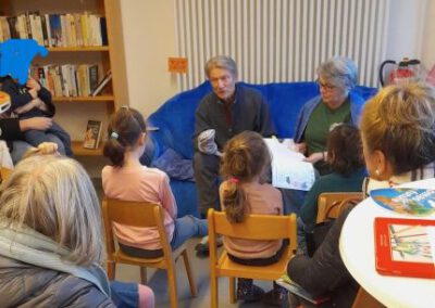 Zwei Erwachsene sitzen auf einer blauen Couch und lesen einer Gruppe von Kindern auf Stühlen in einem gemütlichen Raum mit Bücherregalen und einem Heizkörper im Hintergrund vor. Die Kinder hören aufmerksam zu.