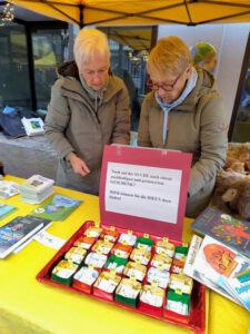 Zwei ältere Frauen stehen an einem gelben Tisch im Freien und ordnen kleine, nummerierte Geschenkkartons. Um ein rotes Schild mit deutschem Text unter einem gelben Baldachin herum sind Bücher und Informationsmaterial ausgestellt.