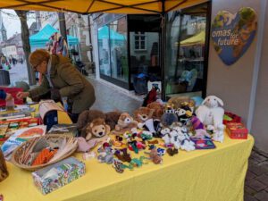 Eine Frau arrangiert auf einem gelben Tisch an einem Marktstand im Freien Plüschtiere, Spielzeug und Handwerksartikel. Hinter ihr steht an einer Wand ein herzförmiges Schild mit der Aufschrift "omas for future". Im Hintergrund sind andere Stände und Menschen zu sehen.