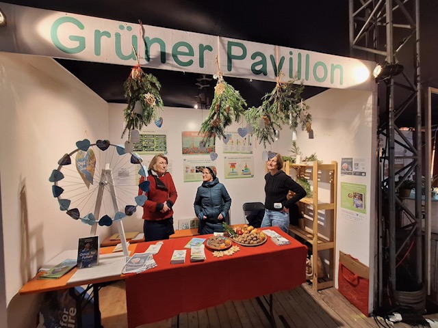 Drei Frauen stehen und unterhalten sich an einem Stand mit der Aufschrift Grüner Pavillon in Leipzig, der mit hängendem Grün dekoriert ist. Der Stand enthält Broschüren, ein Glücksrad und Snacks, die für Umwelt- oder Gemeinschaftsaktivitäten werben.