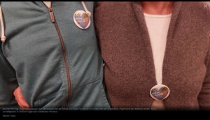 Zwei Personen stehen nebeneinander und tragen Jacken, an deren Brust jeweils ein runder Knopf mit der Aufschrift omas für Zukunft befestigt ist. Die Köpfe sind aus dem Bild herausgeschnitten.