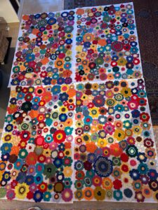 Haekeln Muenchen 03 2026 ©O4F - Omas for Future Vier große Tafeln mit bunten, gehäkelten Blumenmotiven sind nebeneinander auf dem Boden ausgelegt und bilden ein lebendiges und kompliziertes Patchwork-Muster, das auf subtile Weise die Themen Natur und Ökozid widerspiegelt. Jedes Feld weist eine Vielzahl von Formen und Farben auf.