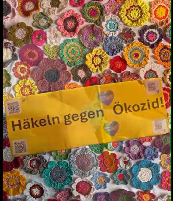 Eine bunte Häkeldecke mit verschiedenen Blumenmustern, gekrönt von einem gelben Schild mit der Aufschrift Häkeln gegen Ökozid! (Häkeln gegen Ökozid) auf Deutsch.