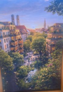 Muenchen Stadt fuer alle 2026 ©O4F - Omas for Future Eine lebendige Stadtlandschaft bei Sonnenuntergang zeigt üppiges Grün, Wohnhäuser mit Balkonen, Menschen, die sich auf einem Platz versammeln, und zwei Kirchtürme im Hintergrund unter einem bunten Himmel.