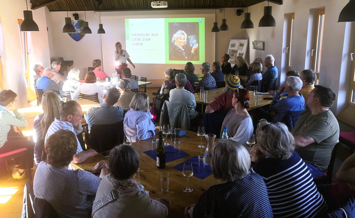 Eine Gruppe von Menschen sitzt an Tischen in einem hellen Raum und beobachtet aufmerksam eine Frau, die einen Vortrag hält, dessen Folien auf eine Leinwand an der Stirnseite projiziert werden. Gläser und Flaschen stehen auf den Tischen.