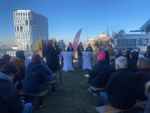 Pressekonferenz Stadt fuer alle Muenchen 2026 ©O4F - Omas for Future Eine Gruppe von Menschen sitzt auf Bänken im Freien, gegenüber von drei Rednern auf Podien. Die Veranstaltung