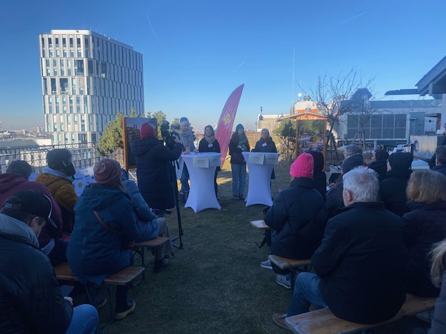 Die Menschen sitzen auf Bänken im Freien und blicken auf drei Redner, die auf einer Dachterrasse auf einem Podium stehen. Die Veranstaltung findet bei Tageslicht und strahlend blauem Himmel statt, und im Hintergrund ist ein modernes Gebäude zu sehen.