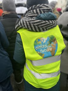 Pruefdemo Muenchen 2026 2 ©O4F - Omas for Future Eine Person, die eine leuchtend gelbe Weste mit einer herzförmigen Weltkugel und der Aufschrift omas for future trägt, steht inmitten einer Menschenmenge auf der Prüf-Demo, von hinten gesehen. Die Person trägt außerdem eine graue Strickmütze und einen gestreiften Schal.
