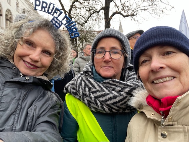 Drei Frauen lächeln bei einem Treffen im Freien bei kaltem Wetter, sie tragen Jacken und Schals. Eine Frau hat lockiges graues Haar und trägt ein Stirnband mit der Aufschrift PRUITTIG. Im Hintergrund sind Menschen und blattlose Bäume zu sehen.