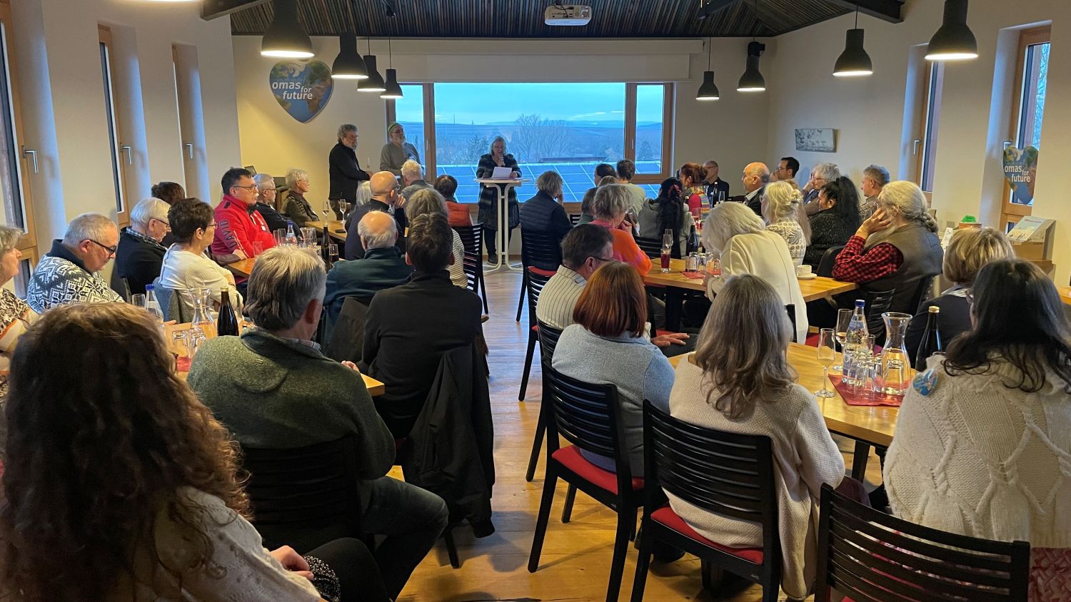 Eine Gruppe von Menschen sitzt an Tischen in einem hellen Raum und hört aufmerksam einem Redner zu, der an einem Podium vor großen Fenstern mit Blick ins Freie sitzt. Auf den Tischen stehen Flaschen und Gläser, die eine zwanglose, gemeinschaftliche Atmosphäre schaffen.