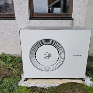 Ein Bosch Wärmepumpe-Außengerät ist neben einer Hauswand auf einer Betonplatte installiert, die von Gras und Erde umgeben ist. Das Gerät ist mit einem runden Lüftungsschlitz und dem Bosch-Logo auf der Vorderseite versehen und liefert erneuerbare Energie für Ihr Haus.