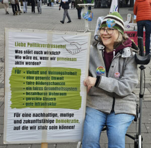 Eine ältere Frau mit Brille und Mütze sitzt in einem Rollstuhl und lächelt neben einem Protestschild. Auf dem Schild stehen Forderungen nach Demokratie, Vielfalt, Umwelt, gerechter Bildung, bezahlbarem Wohnraum und besserer Infrastruktur.