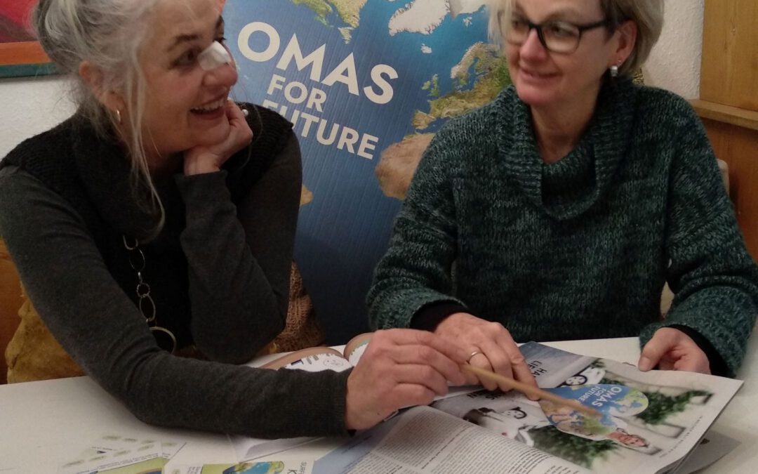 Zwei ältere Frauen sitzen an einem Tisch, lächeln und unterhalten sich, während sie sich Zeitschriften und Broschüren ansehen. Hinter ihnen steht ein Schild mit der Aufschrift OMAS FOR FUTURE. Die Atmosphäre ist freundlich und entspannt.