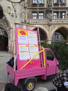 Stadt fuer Alle Forderungen 02 2026 ©O4F - Omas for Future Ein kleiner rosafarbener dreirädriger Lastwagen ist vor einem historischen Steingebäude geparkt. Er trägt eine große rosafarbene Anzeigetafel mit gelber und weißer Schrift, auf der