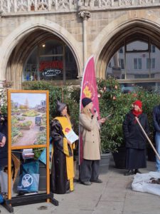 Stadt fuer Alle KI Bild 02 2026 ©O4F - Omas for Future Eine Gruppe von Menschen steht vor einem historischen Gebäude neben einer bebilderten Gartenausstellung und präsentiert ihre Forderungen. Eine Person spricht in ein Mikrofon, andere tragen Kostüme - ein gelbes Gewand, einen roten Hut - und eine lila