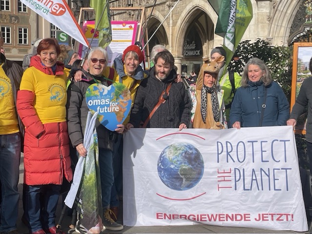 Eine Gruppe von Menschen steht im Freien und hält ein "Protect the Planet"-Transparent und Forderungsschilder, einige tragen gelbe Hemden und einer ein Hirschkostüm, bei einem Klimaprotest. Die Sonne scheint und im Hintergrund sind Gebäude in der Stadt für Alle zu sehen.