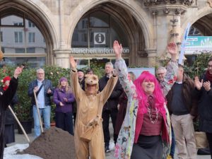 Stadt fuer Alle Tiere 02 2026 ©O4F - Omas for Future Eine Gruppe von Menschen steht vor einem historischen Gebäude. Im Vordergrund feiern zwei Personen mit erhobenen Armen - eine in einem Kamelkostüm, die andere mit einer leuchtend rosa Perücke - während andere zusehen und lächeln, um ihre Unterstützung für