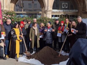 Uebergabe Forderungen Muenchen 02 2026 ©O4F - Omas for Future Eine Gruppe von Menschen steht im Freien vor der München Information, versammelt um einen Erdhügel, der mit einer weißen Plane abgedeckt ist. Einige halten Zettel mit Forderungen in der Hand, und einige tragen Kostüme, während das Sonnenlicht die Szene beleuchtet.