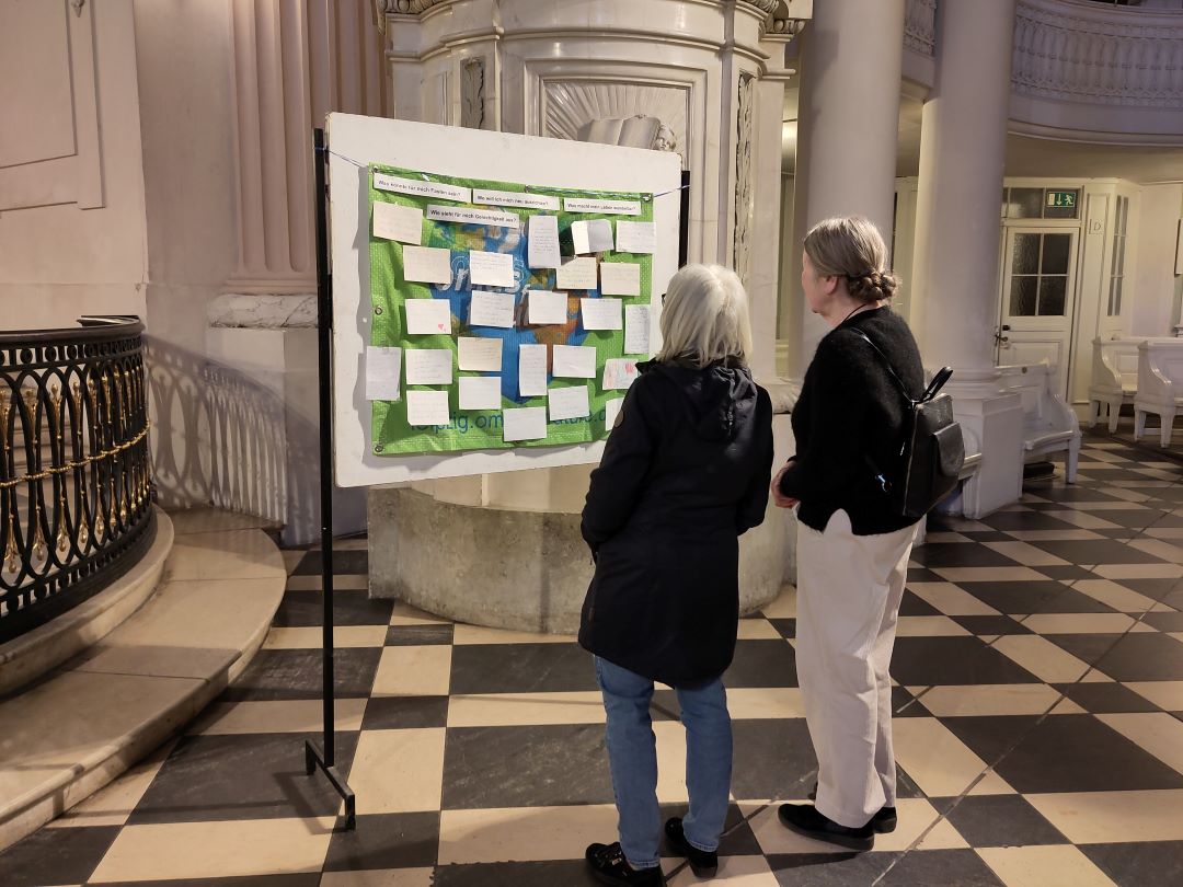 Zwei Personen stehen in einem Innenraum vor einer großen Schautafel, die mit verschiedenen Notizen und Papieren bedeckt ist, und lesen deren Inhalt. Die Kulisse besteht aus Säulen, schachbrettartigen Bodenfliesen und verschnörkelten architektonischen Details.