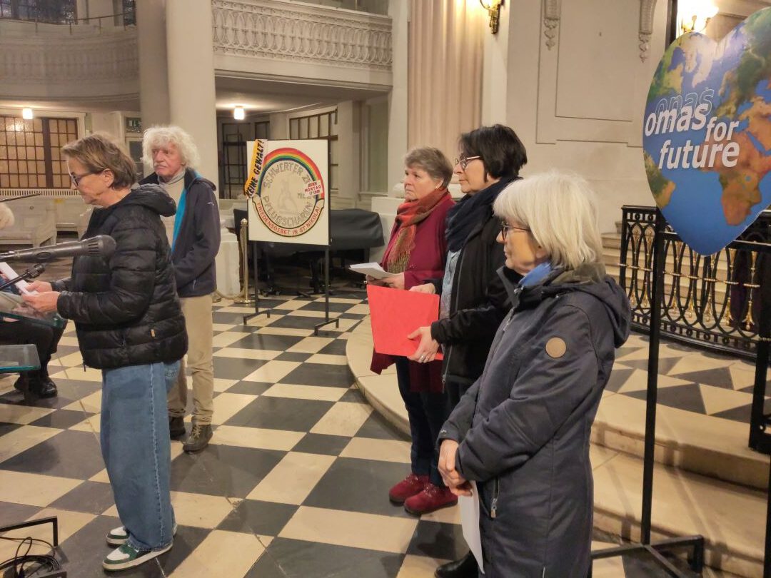 Eine Gruppe älterer Menschen steht drinnen, einige mit Papieren in der Hand, neben einem blauen Schild mit der Aufschrift omas for future. Die Umgebung wirkt förmlich, mit kariertem Boden und verschnörkelten Geländern.