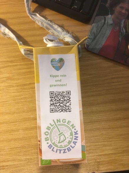 Ein Schlüsselband mit einer Plakette auf einer Holzoberfläche. Auf dem Anstecker steht "Kippe rein und gewinne!" mit einem QR-Code darunter und einem grünen Böblingen Blitzblank-Logo am unteren Rand. Ein Teil eines Mauspads ist sichtbar.