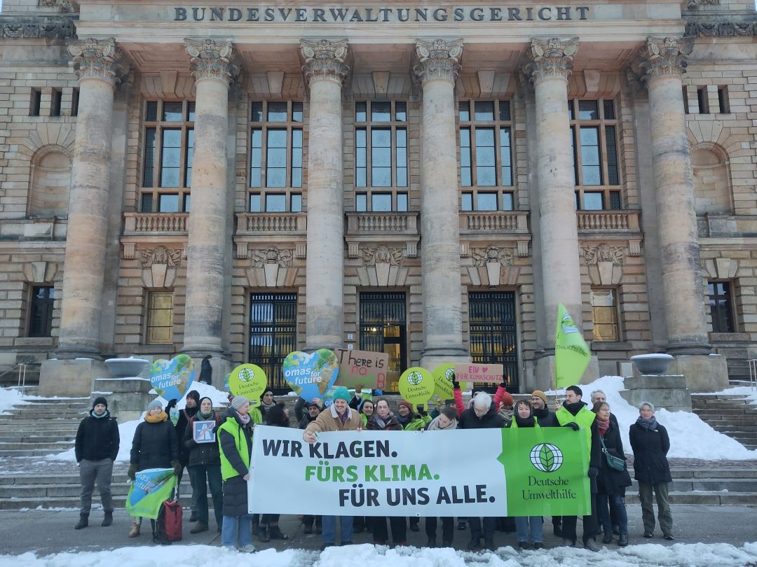 Eine Gruppe von Menschen steht vor dem Gebäude des Bundesverwaltungsgerichts und hält ein Transparent mit der Aufschrift "Wir klagen. Fürs Klima. Für uns alle." bei Schneetreiben, mit Klimaschildern und Fahnen.