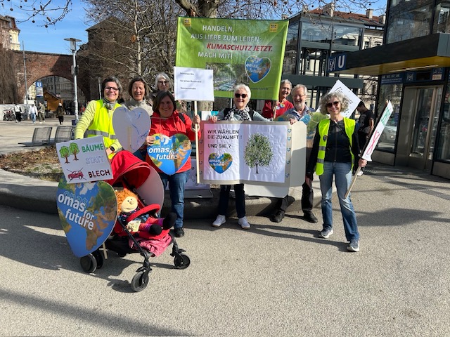 Eine Gruppe von Senioren und Seniorinnen steht in München im Freien und hält Klimaschutz-Protestschilder, darunter "Omas for Future". Eine Person hält einen Kinderwagen mit einem Teddybär, und die fröhliche Gruppe genießt den sonnigen Himmel.