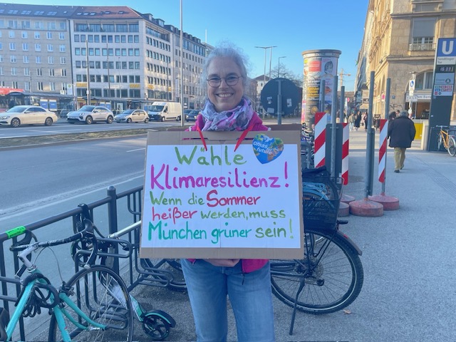 Eine ältere Frau mit grauen Haaren und Brille steht auf einem Bürgersteig in der Stadt und hält ein handgemaltes Klimaschutz-Schild in die Höhe, das für Klimaresilienz und grünere Sommer in München wirbt. Im Hintergrund sind Fahrräder und Gebäude zu sehen.