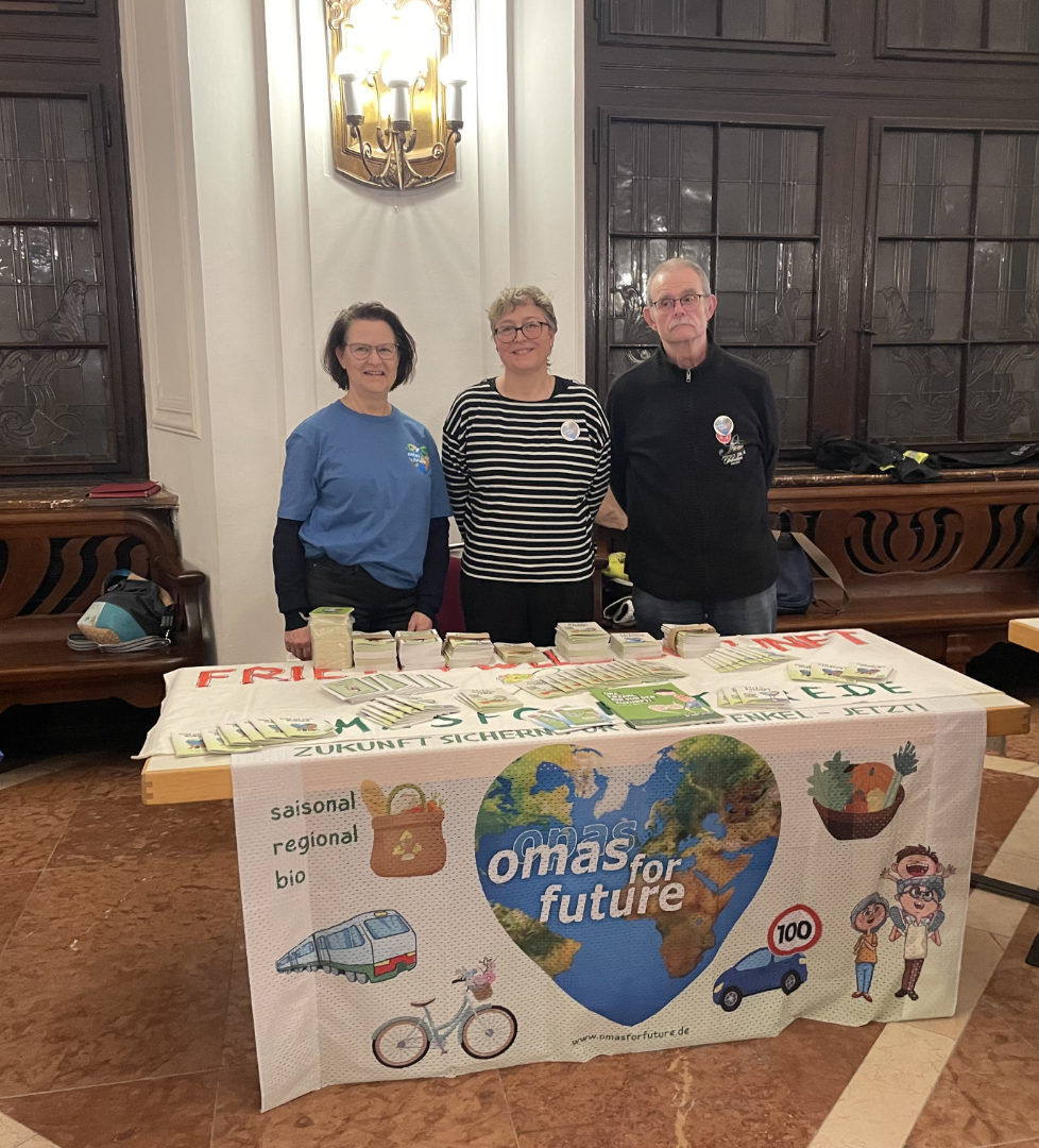 Drei Personen stehen hinter einem Tisch mit einem farbenfrohen "Omas for Future"-Transparent, das mit Umweltillustrationen, Broschüren und Gläsern geschmückt ist, in einem geräumigen Innenbereich.