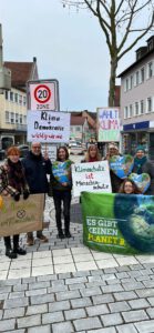 8d59e7b2 c9b5 4994 bd01 ac1f8847ef0e - Omas for Future Eine Gruppe von Menschen steht und kniet auf einer Kopfsteinpflasterstraße und hält Schilder zum Klimaschutz, zur Demokratie und zum Umweltschutz in die Höhe. Im Hintergrund sind Gebäude, ein Schild mit einer 20 km/h-Zone und ein blattloser Baum zu sehen.