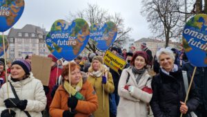 Anti Sellner Demo Dez24 Augsburg©O4F - Omas for Future Eine Gruppe älterer Frauen hält bei einem Protest herzförmige Schilder mit der Aufschrift Omas for Future. Sie tragen Winterkleidung und stehen im Freien mit anderen Demonstranten und Bäumen im Hintergrund. Es sind Mitglieder der Regionalgruppe Augsburg