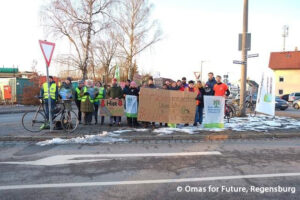 Eine Gruppe von Menschen steht auf einem Bürgersteig an einer Straße und hält Schilder und Transparente in deutscher Sprache. Einige tragen Sicherheitswesten, und um sie herum stehen Fahrräder und Schneeflecken. Im Hintergrund sind Fahnen und Gebäude zu sehen.