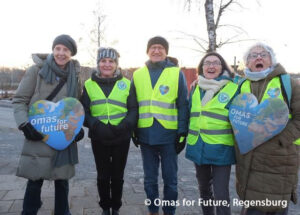 Fünf ältere Erwachsene in Winterkleidung und gelben Sicherheitswesten stehen im Freien, lächeln und halten herzförmige Schilder mit der Aufschrift Omas for Future. Im Hintergrund sind Bäume und ein kalter, sonniger Tag zu sehen.