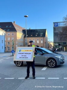 Eine Person steht vor einem grauen Auto auf einer Straße in der Stadt und hält ein großes Schild in der Hand, das dazu auffordert, für die Zukunft mit Fahrrädern, Bussen und Zügen zu stimmen. Im Hintergrund sind Gebäude und ein Parkplatzschild zu sehen.