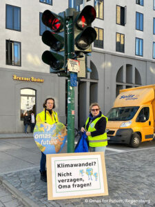 Zwei Frauen in gelben Westen stehen an einer Ampel und halten ein herzförmiges Schild mit der Aufschrift Omas for Future. Auf einem großen Schild darunter steht: Klimawandel? Nicht verzagen, Oma fragen! In der Nähe ist ein gelber Lieferwagen geparkt.