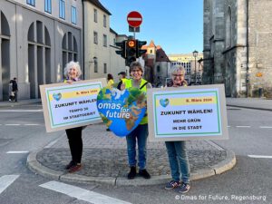 Drei ältere Frauen stehen auf einer Straße und halten Schilder, die zu Umweltschutzmaßnahmen in Regensburg, Deutschland, aufrufen. Sie werben für "Omas for Future" und setzen sich für eine grünere Politik und niedrigere Geschwindigkeitsbegrenzungen in städtischen Gebieten ein.