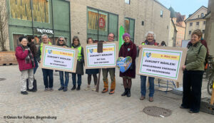Eine Gruppe von Menschen steht im Freien und hält Schilder in deutscher Sprache, die für erneuerbare Energien und nachhaltigen Verkehr in Regensburg werben. Auf den Schildern steht "Zukunft wählen!" ("Zukunft wählen!"). Hinter ihnen sind Gebäude und Geschäfte zu sehen.