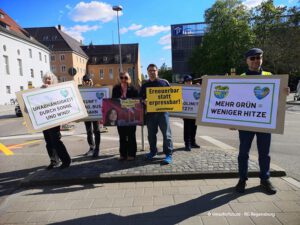 Eine Gruppe von Menschen steht im Freien und hält Protestschilder in deutscher Sprache über erneuerbare Energien und Klimaschutz. Der Himmel ist klar und im Hintergrund sind mehrere Gebäude zu sehen.