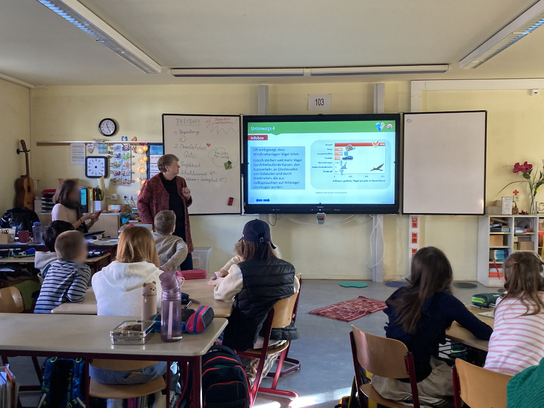 Kinderquiz in 39. Oberschule©O4F - Omas for Future Ein Klassenzimmer mit Schülern, die an Tischen sitzen und einem Lehrer gegenübersitzen, der eine Unterrichtsstunde auf einem großen digitalen Bildschirm präsentiert.