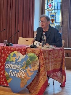 Eine Person spricht an einem Tisch, der mit einem rot gemusterten Tuch und einem herzförmigen Schild mit der Aufschrift "omas" für "Zukunft" bedeckt ist; vor ihr stehen Mikrofone, eine Wasserflasche und Bücher. Im Hintergrund ist ein Buntglasfenster zu sehen.