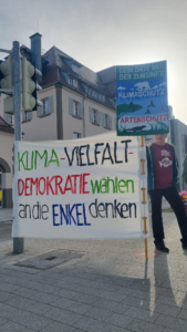 Eine Person steht im Freien und hält ein großes Transparent mit der Aufschrift "Klima - Vielfalt - Demokratie wählen, an die Enkel denken". Außerdem hält sie ein Schild, auf dem für Klimaschutz und Artenvielfalt geworben wird. Im Hintergrund sind Gebäude zu sehen.