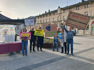 Sechs Personen stehen im Freien und halten bunte Schilder in die Höhe, auf denen sie für Klimaschutz und Wahlen werben. Sie stehen neben einem Informationsstand mit Plakaten, vor einem Gebäude mit großen Fenstern und roten Schildern.